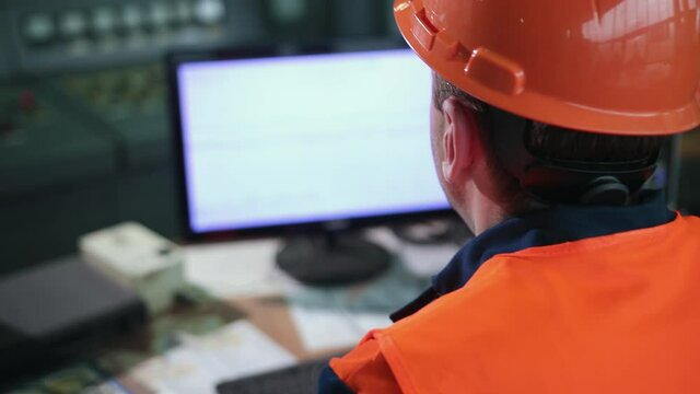 Inside heavy industry, factory male industrial engineer works on personal computer. Industrial engineer in hard hat wearing safety jacket uses computer. Works at heavy industry manufacturing factory