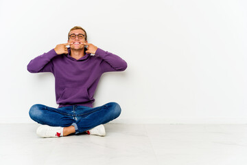Young caucasian man sitting on the floor smiles, pointing fingers at mouth.