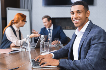 Happy african american businessman looking at camera while typing on laptop with blurred colleagues talking on background.