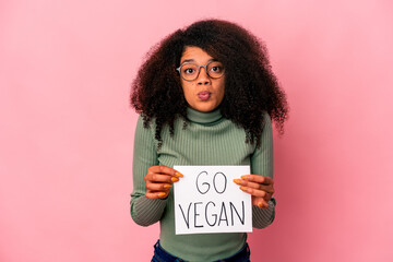 Young african american curly woman holding a go vegan placard shrugs shoulders and open eyes...