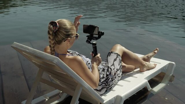 Woman shooting on handheld film gimbal stabilization for smartphone. Girl rest, relax, lie sunbed on pier in sunglasses, make selfie. Lady blogger broadcast video blogging vlogging. Take photo video.