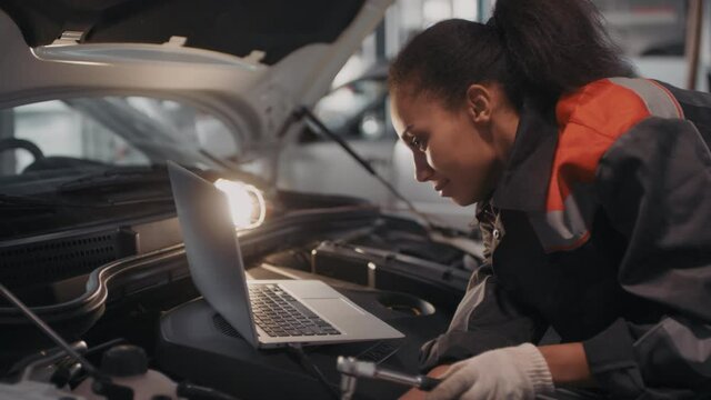 Handheld shot of cheerful black female auto mechanic in gloves typing on laptop lying under hood of car and making repairs with tool