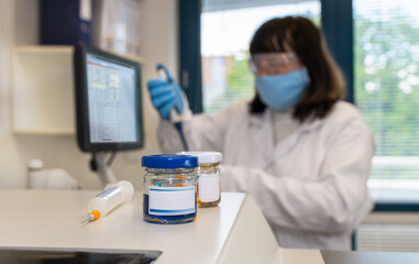 Clinical laboratory scientist in protective face mask and glasses holding pipette. Close-up of glass vials and a syringe on lab analyzer and female medical technologist working with computer software.