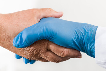 Helping hand of health professional holding old wrinkly palm on white background. Detail of safe handshake of doctor in blue protective glove and senior patient. Elderly care concept in pandemic time.