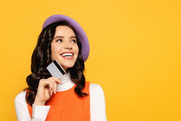  brunette woman in beret looking up while holding credit card isolated on yellow