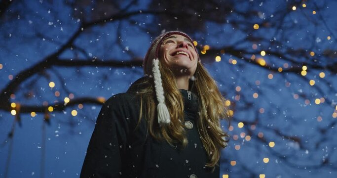 Cinematic shot of carefree happy smiling young woman wearing warm hat is excited and amazed with Christmas lights on the tree in a festive winter evening.Concept of winter holidays, freedom, happiness
