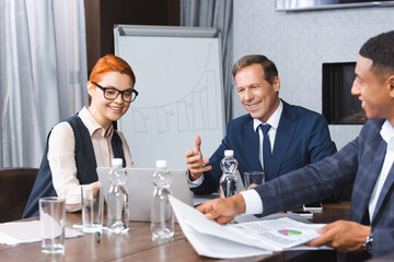 Smiling businesspeople looking at laptop while sitting at workplace near african american businessman with documents.