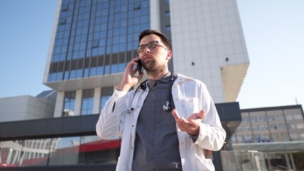 Young Caucasian doctor in white medical coat consults patient on cell phone during break outside hospital. Medical worker talking on mobile phone near clinic. Physician in uniform talk on phone