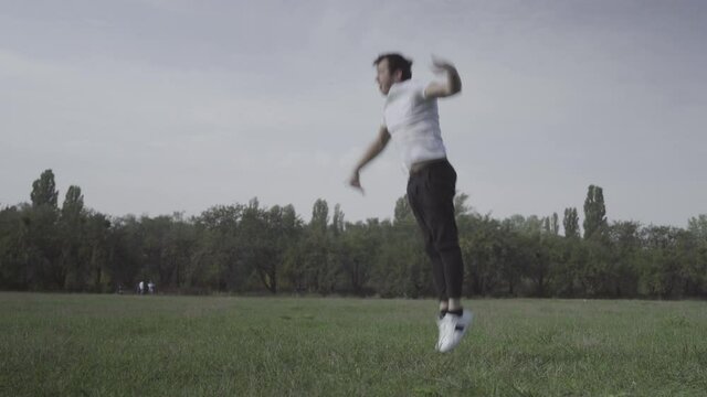 Wide shot of young break dancer dancing on green summer meadow. Portrait of confident brunette Caucasian man training dance outdoors. Lifestyle and hobby concept.