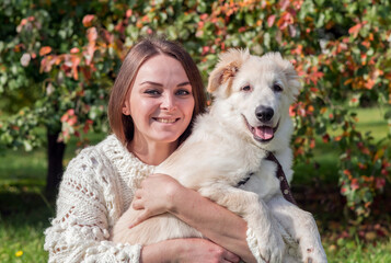 Beautiful caucasian woman in white sweater holds and strokes labrador puppy sitting on green grass in autumn city park on a sunny day during dog walking. The puppy is about 5 months old. Pets theme.