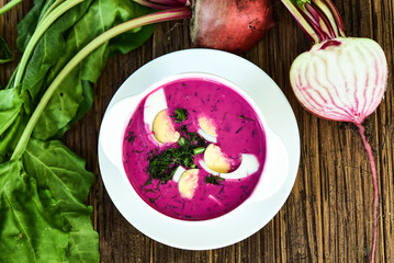 Cold beetroot soup, cold served with the addition of cucumbers, radishes, boiled eggs and fresh herbs, dill, green onions on a wooden table, traditiona Chilled polish cold soup
