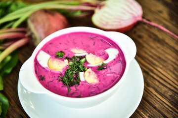 Cold beetroot soup, cold served with the addition of cucumbers, radishes, boiled eggs and fresh herbs, dill, green onions on a wooden table, traditional chilled polish cold soup