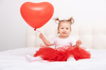 Fototapeta na wymiar cute baby girl holds heart balloon sits on a white bedding.
