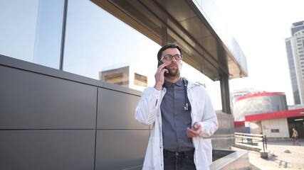 Fototapeta premium MS doctor in white coat talking on mobile phone front of clinic. Man medic outside speaking on cell phone. Medicine healthcare professional theme. Physician discussing with patient during phone talk