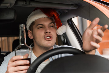 displeased, drunk man in santa hat gesturing while sitting at steering wheel with bottle of whiskey, blurred foreground.