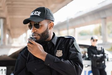 african american police officer talking on radio set near policewoman on blurred background outdoors.