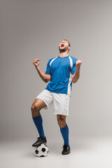 Excited sportsman in sportswear showing yeah gesture near football on grey background