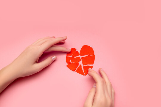 Female hands gluing together broken heart, pieces of torn red valentine on pink background. The concept of disappointment in love, salvation of relationships. Close-up photo