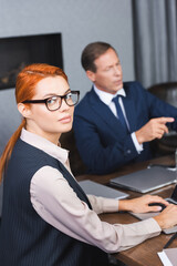 Fototapeta na wymiar Redhead businesswoman in eyeglasses looking at camera while typing on laptop with blurred investor on background.