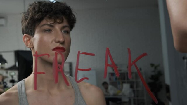 Angry trans man stares at the word FREAK on the mirror. Androgynous guy with makeup looks at his reflection. Gender identity and social label concept 4k