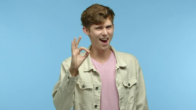 Stylish young man in casual clothes, saying yes, oh hear and making okay gesture, nod in approval, praise something good, standing over blue background