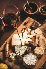 Assortment of snacks and appetizers for red wine. Various cheeses on sticks with bull, crackers, dry fruit and glasses of red wine on wooden rustic board over rusty brown background, selective focus