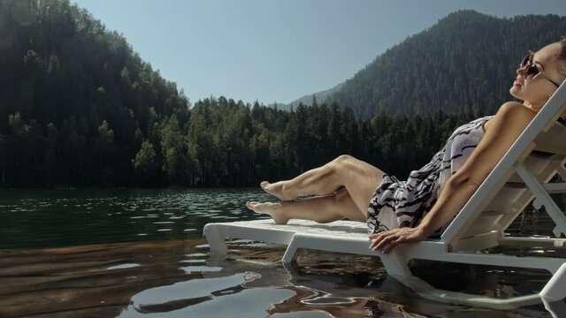 Woman lie on a sunbed in sunglasses and a boho silk shawl. Girl rest on a flood wood underwater pier. The pavement is covered with water in the lake. In the background are mountain and a forest.