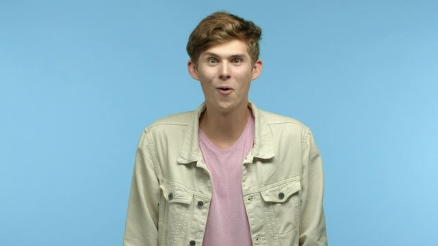 Slow motion of handsome young man checking out cool promo offer, raising eyebrows and looking surprised, smiling and nodding to praise something awesome, blue background