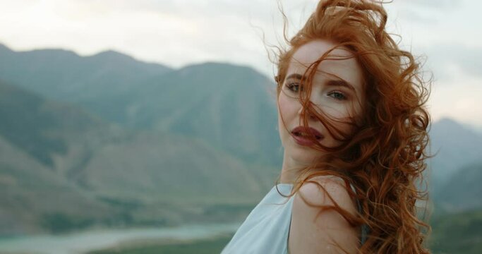 Beautiful female model on top of a mountain posing for photoshoot, reaching her hand out to camera while wind blows her dress and red hair - freedom, tranquility 4k footage
