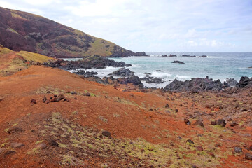 Small beach on the coast of Easter Island, covered by red volcanic dust, against a blue sky with white clouds.