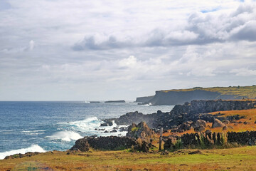 View of the coast of Easter Island, covered by red volcanic dust and green vegetation, against a blue sky with white clouds.