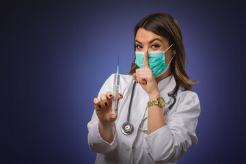 Young female doctor with stethoscope and face mask holding COVID-19 vaccine making hush gesture....