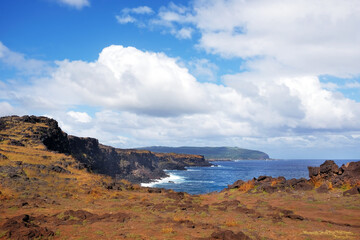 View of the coast of Easter Island, covered by red volcanic dust and green vegetation, against a blue sky with white clouds.