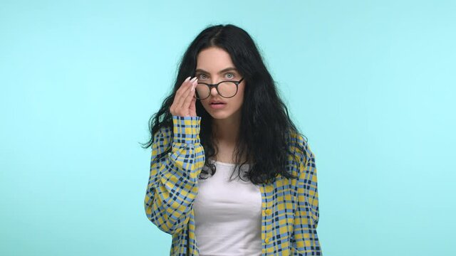 Beautiful young woman with long dark hair looks confused, take-off glasses and blink puzzled, stare at camera with disbelief, cant understand what happen, standing over blue background