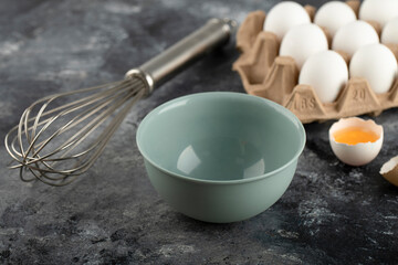 White eggs in carton container, bowl and whisker on marble background