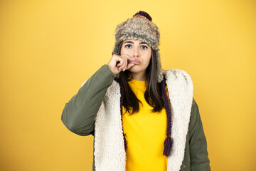 Young beautiful woman wearing a hat and a green winter coat over yellow background with mouth and lips shut as zip with fingers. Secret and silent, taboo talking