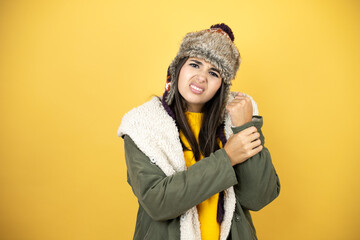 Young beautiful woman wearing a hat and a green winter coat over yellow background suffering pain...