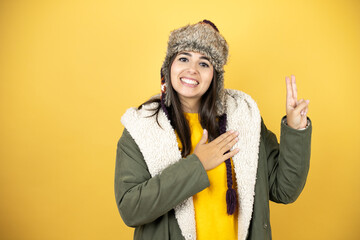 Young beautiful woman wearing a hat and a green winter coat over yellow background smiling swearing...