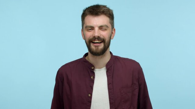 Carefree young man with beard, wearing casual red shirt and t-shirt, laughing out loud and smiling carefree, touching chest while chuckle over funny thing, looking at camera, blue background