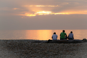 Young people sit by the sea and watch the sunset. Quiet, calm, windless evening on the beach. Pink, colorful sky.