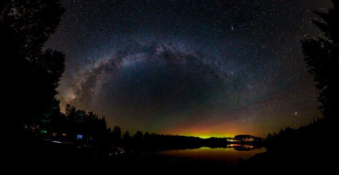 A dramatic panorama of the Milky Way Galaxy and the Northern Lights (Aurora Borealis) high in the night sky above a lake at Algonquin Park, Ontario, Canada