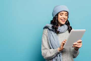  brunette woman in hat and scarf using digital tablet isolated on blue