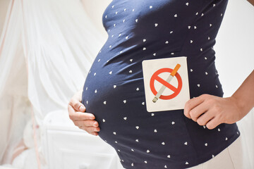 Pregnant woman holding no smoking sign label on belly in baby bedroom. Unhealthy concept.