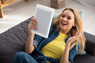  blonde woman with waving hand looking at digital tablet while lying on couch on blurred background