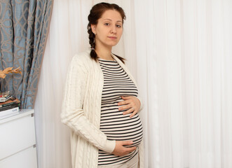 Young beautiful pregnant woman holding her belly and standing near window at home. Concept of pregnancy preparation and expectation, gynecology. Mother with big belly, hands over tummy.