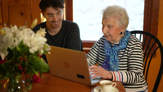 Happy Senior Elderly Woman Sitting With Teen Grandson Learning To Use Laptop Computer In Dining Room.