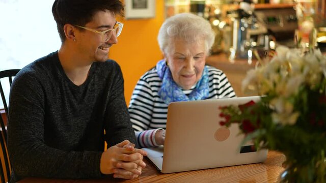 Senior Elderly Woman And Teen Grandson Happy And Smiling Learning On Laptop Computer In Dining Room.