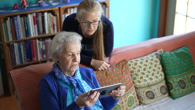 High angle view of mature woman leans over to help elderly senior woman with tablet computer in a living room.