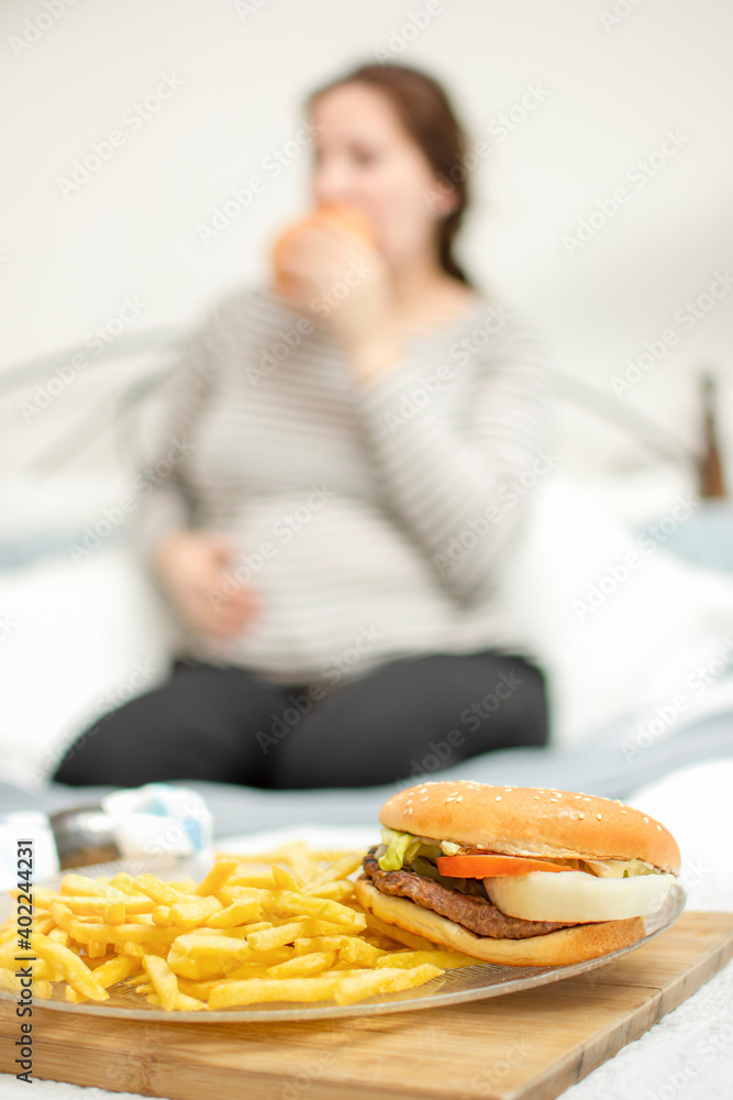 Wall mural young hungry pregnant woman eating a hamburger and fried potatoes. Concept of unhealthy nutrition during pregnancy, Mother waiting baby. Close-up of belly and junk food (fast food). 