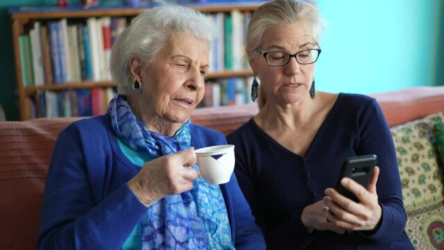 Elderly senior woman and mature daughter drink tea looking at smartphone in living room.
